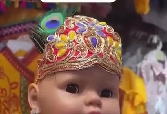 A baby doll wearing a gold and red embroidered crown topped with a peacock feather.