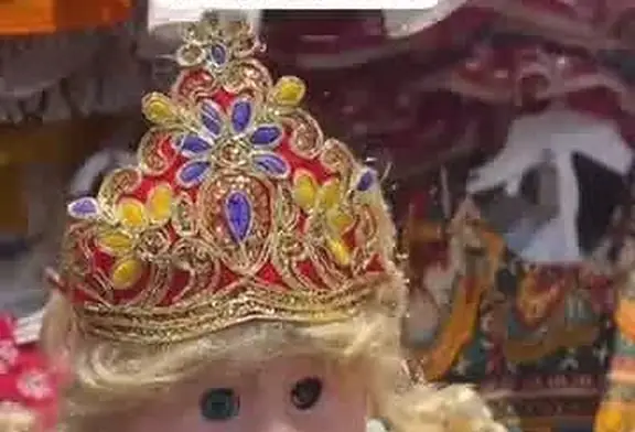 A baby doll in a matching gold and red embroidered crown, worn with a blonde wig.