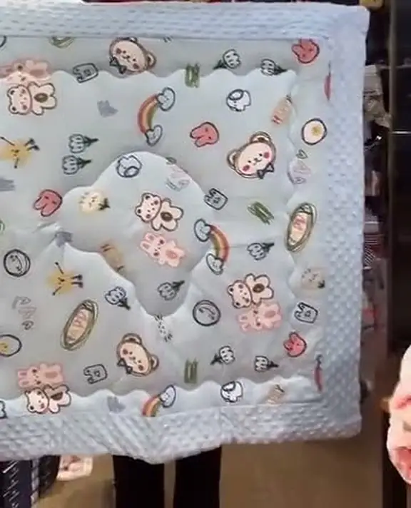 A pale grey quilted muslin blanket printed with bears, rabbits and rainbows, held up in the shop.
