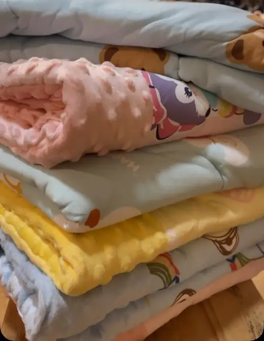 A stack of folded muslin baby blankets in pale blue, pink, mint and yellow, with bear, elephant and rainbow prints.
